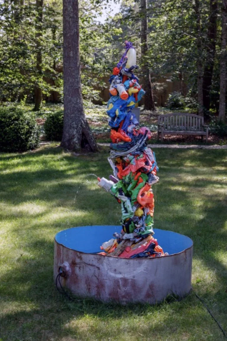 Sculpture made of colorful discarded plastic items stacked in a vertical pile in a park.