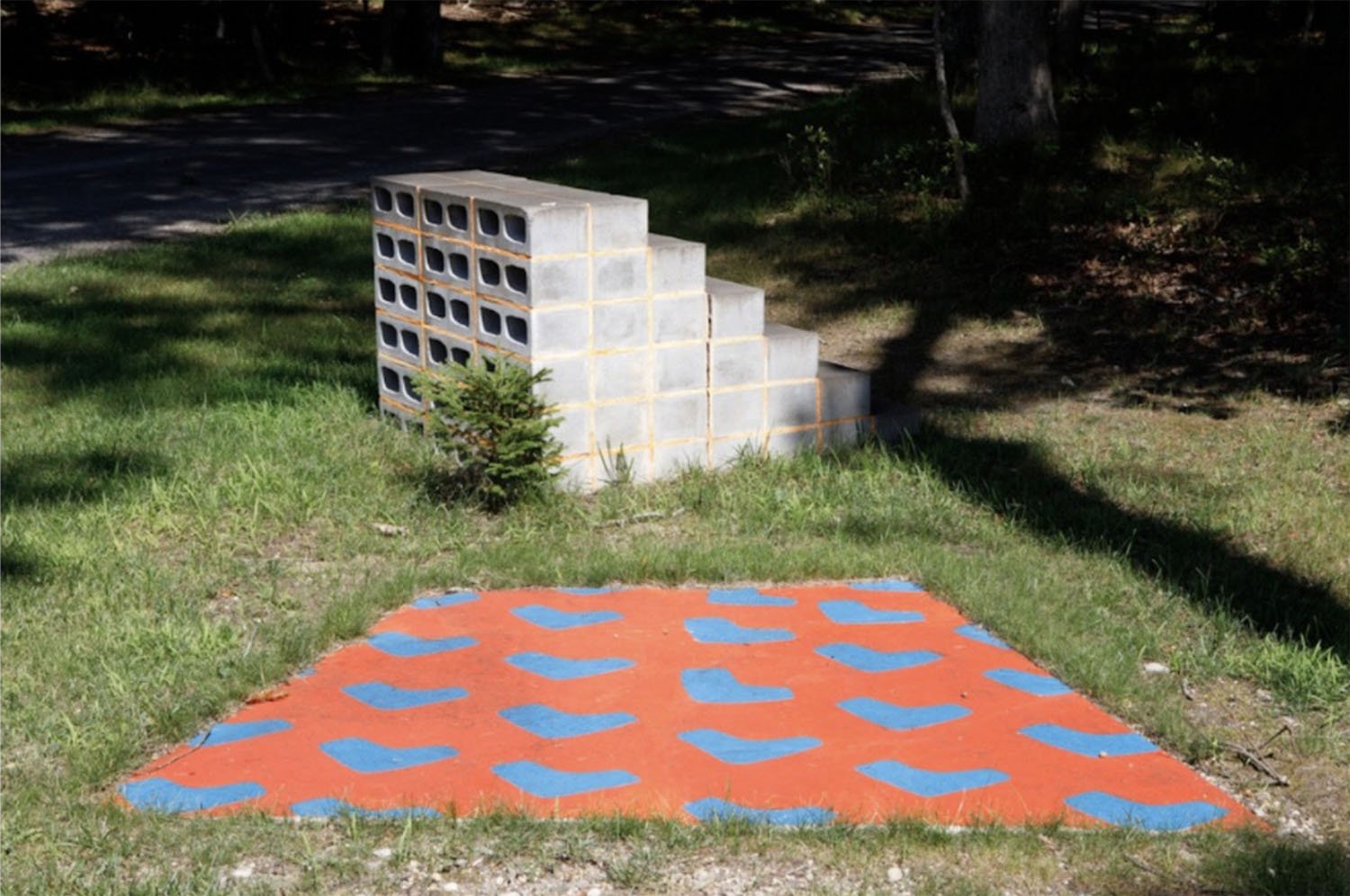 Concrete cinder blocks stacked into a step shape, with a small pine tree in front, on grass. In the foreground, an orange and blue heart pattern painted on the ground.