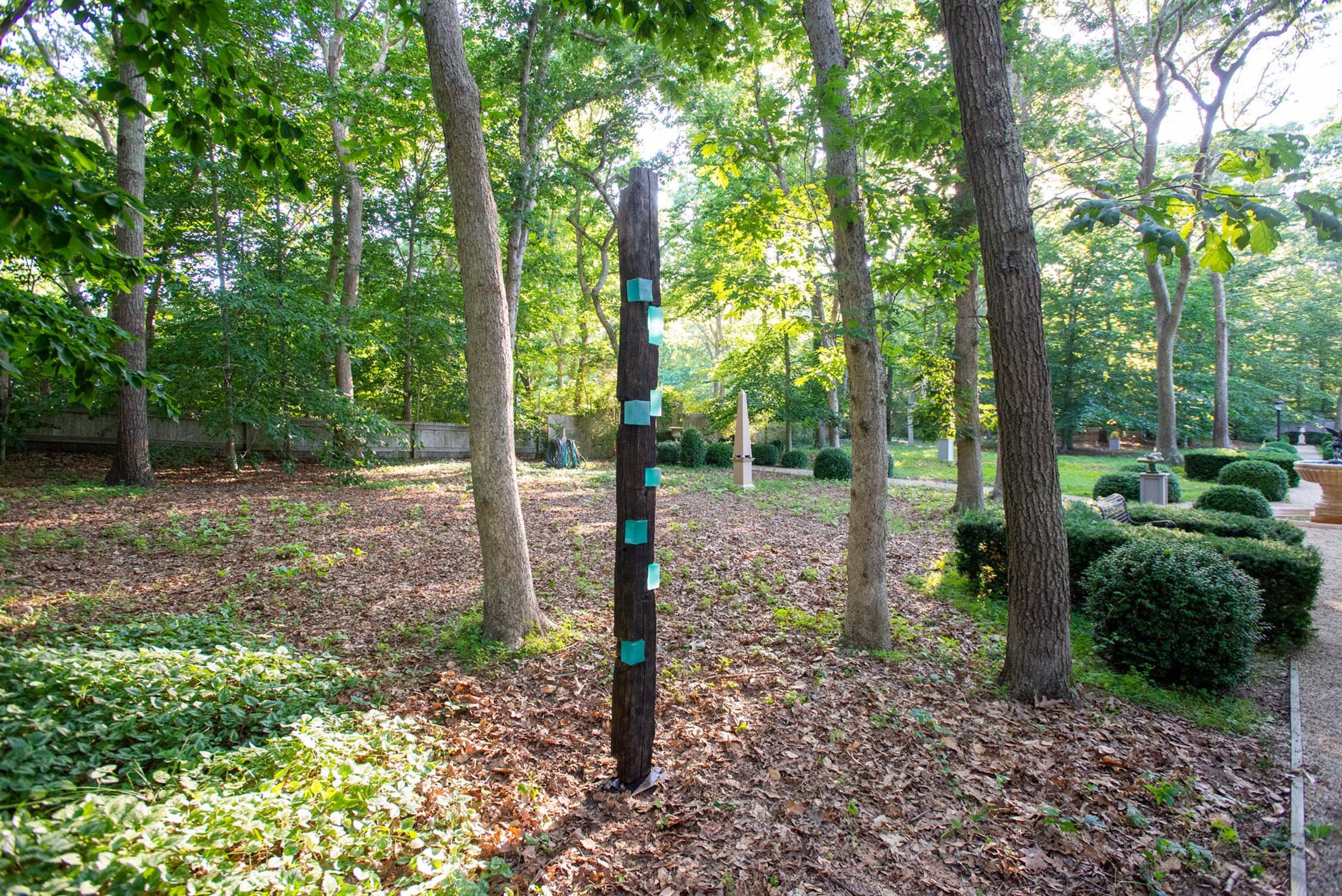 A wooded area of The Leiber Collection Sculpture Garden with a sculptures of weathered wooden post with patches of blue paint and plaques attached. There are tall trees, green bushes, and a gravel path to the right.