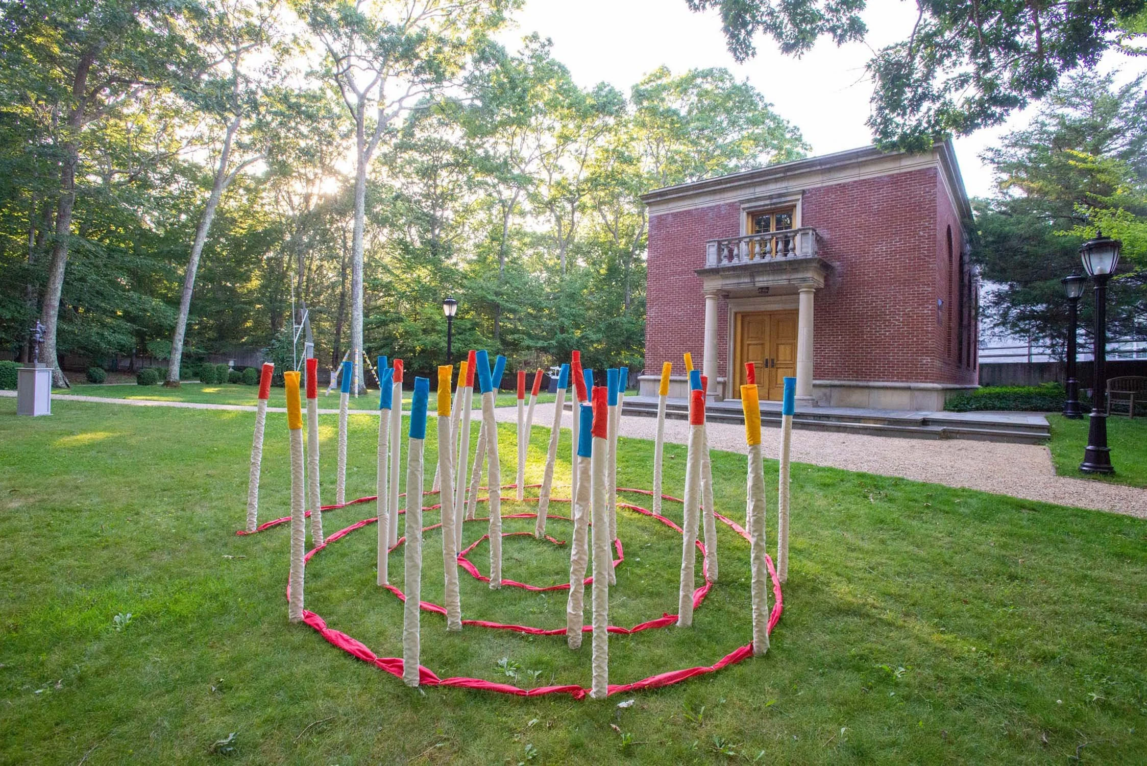 Outdoor scene with a circular array of tall, thin, striped sticks with colored tips, set on a grassy lawn in front of The Leiber Collection museum red brick building with columns and a small balcony, surrounded by trees and lamp posts.