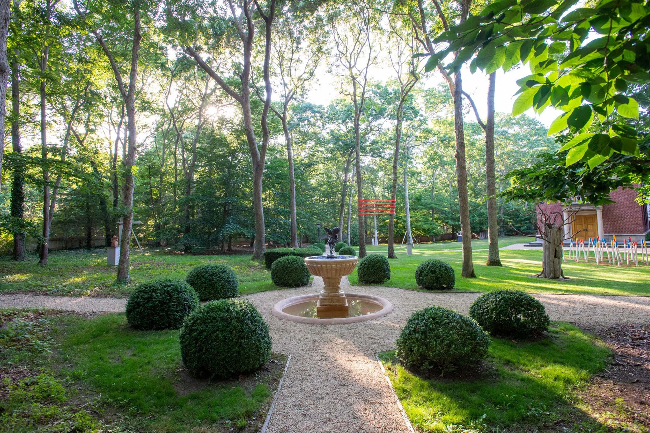 Sunlight filters through trees onto a landscaped garden with a central fountain surrounded by trimmed bushes, a gravel pathway, and a grassy area with scattered trees.