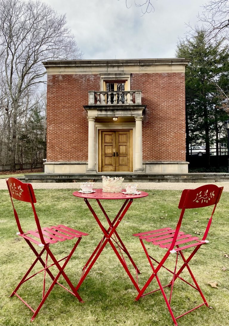 Outdoor scene featuring a small red table with two matching red chairs on green grass, set in front of a historic brick building with wooden double doors, columns, and a small balcony.