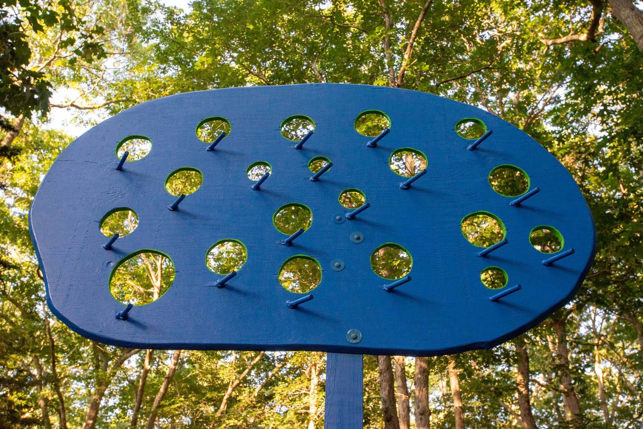 Blue wooden outdoor sculpture with circular cutouts and metal pegs, set against a background of trees with green leaves and sunlight.