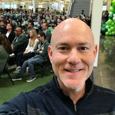 A man taking a selfie at a large indoor event with many seated attendees and green and black balloons in the background.