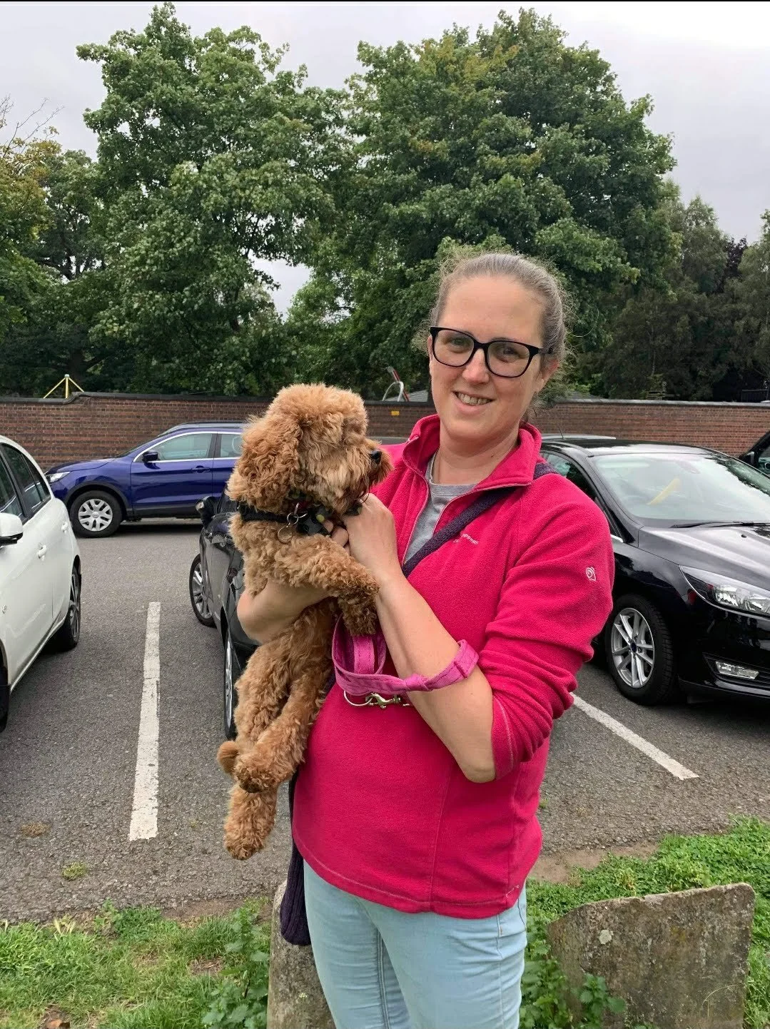 Helen standing holding a brown cavapoo dog.  Helen is smiling, hair tied back, and wearing glasses and a pink jumper.  There are cars and trees in the background.