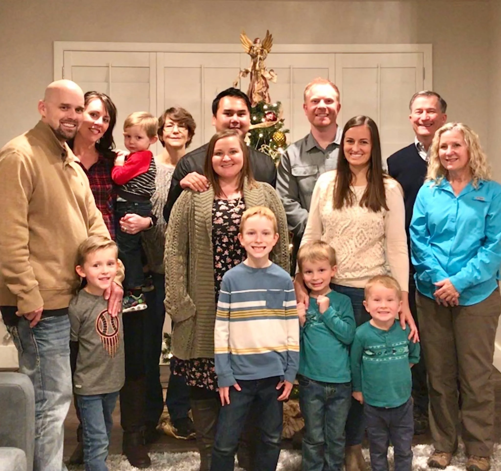Family group photo celebrating Christmas with a decorated tree in the background, including adults and children smiling indoors.