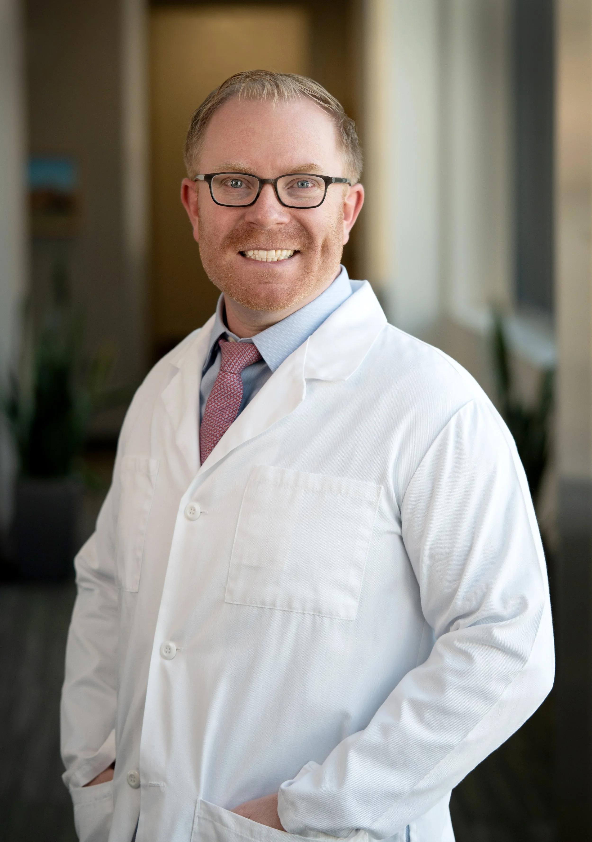A smiling male doctor with glasses, wearing a white lab coat, blue shirt, and pink tie, standing indoors near a window.