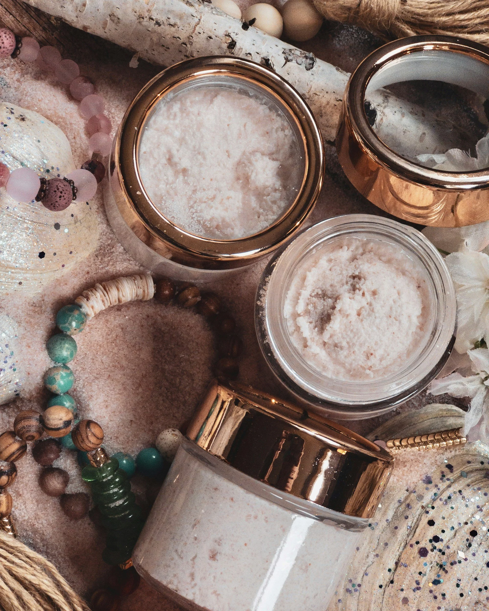 Various jars and containers of bath salts on a pink sandy surface, surrounded by decorative beads, pearls, and glittered ornaments.