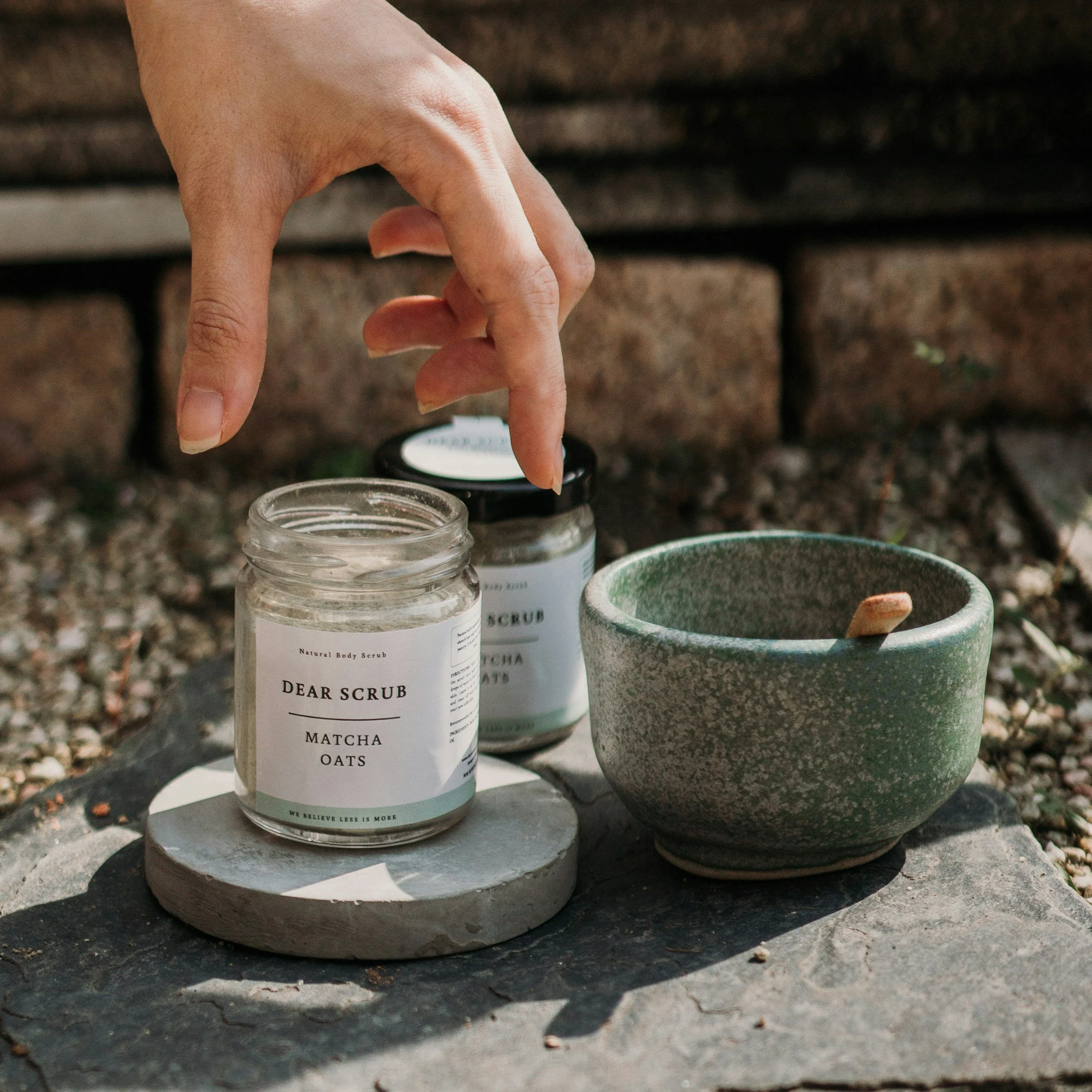 A person’s hand reaching to adjust a small jar labeled 'Dear Scrub Matcha Oats' next to another jar with a black lid and a green ceramic bowl on a rock surface outdoors.
