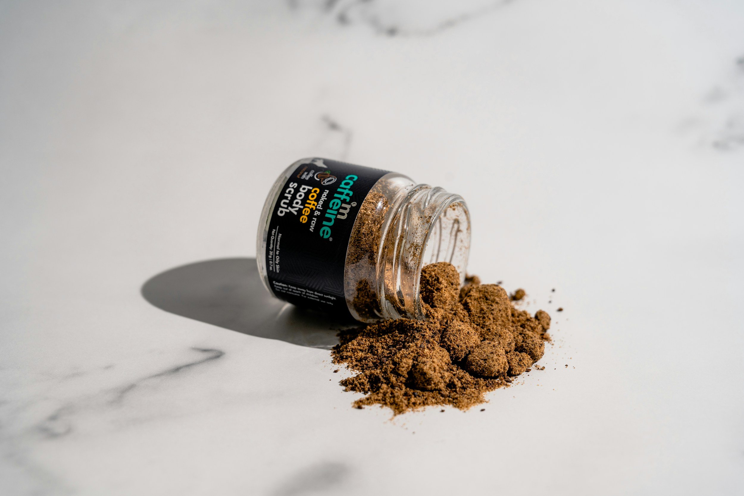 A small glass jar labeled 'caffeine body scrub' from the brand 'Caffeine' tipped over on a white marble surface, with brown coffee scrub spilled out around it.