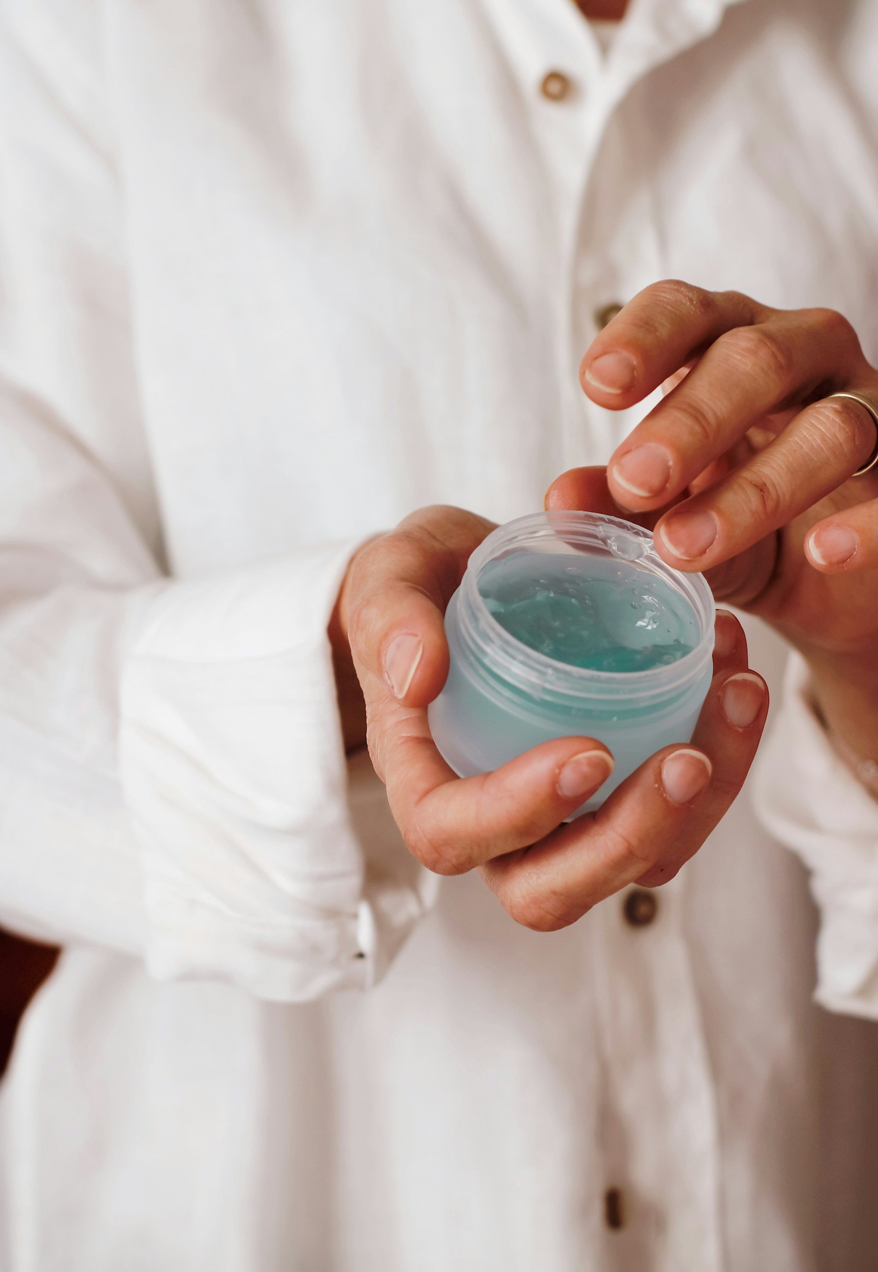 A person in a white shirt holding a small jar of light blue gel or cream, opening it with fingers.