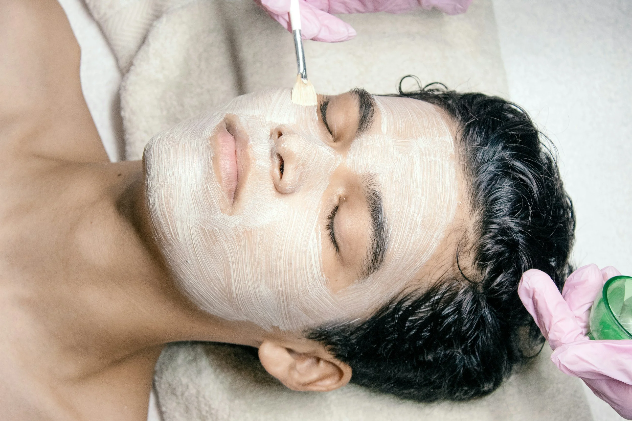 A man is receiving a facial treatment with a mask applied to his face, while lying down with eyes closed.