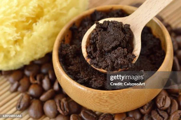 Close-up of a wooden bowl filled with ground coffee, with a wooden spoon scooping some of the coffee, surrounded by coffee beans and topped with a yellow, fluffy substance.