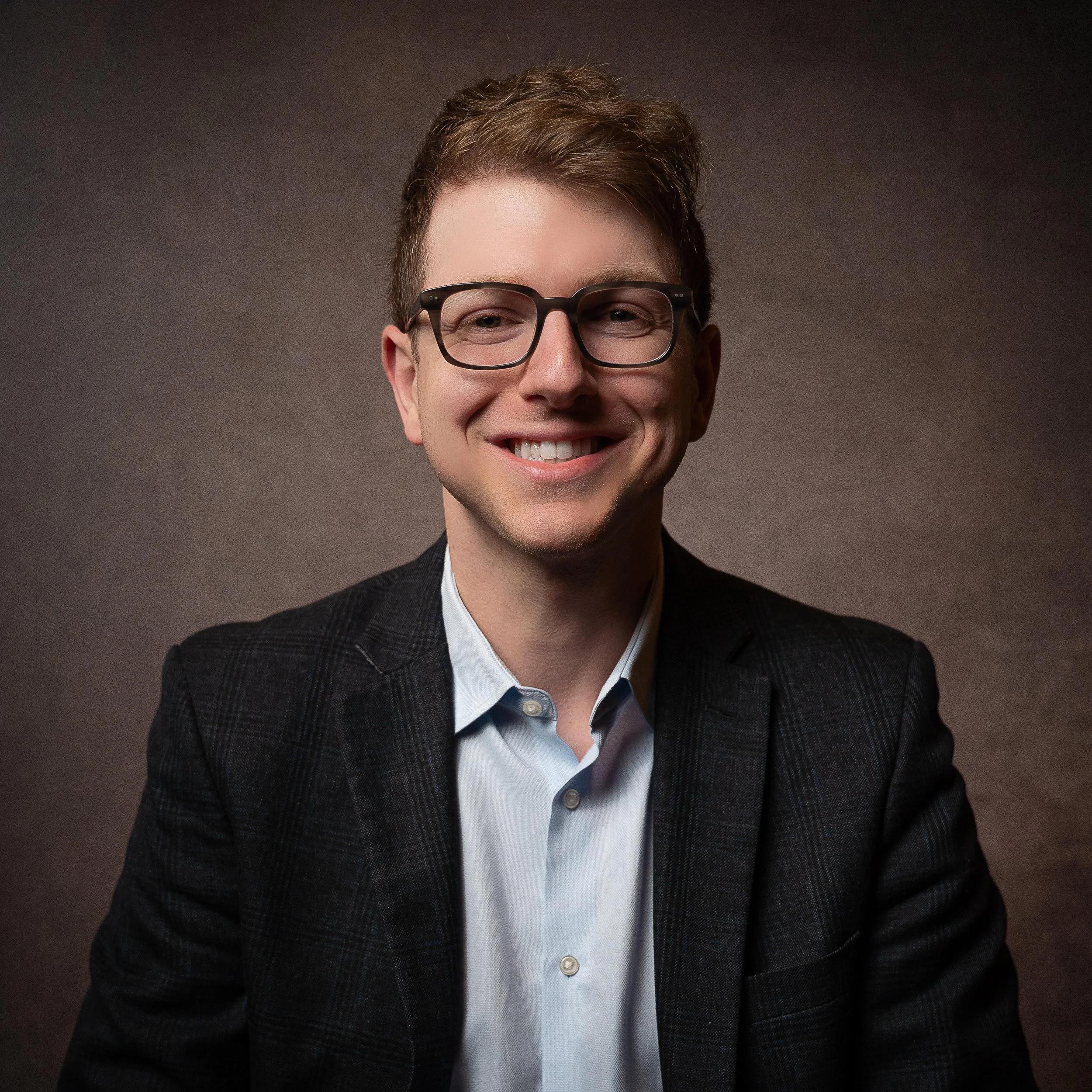 A smiling young man with short brown hair, wearing glasses, a dark blazer, and a light blue collared shirt, posing against a plain, dark background.