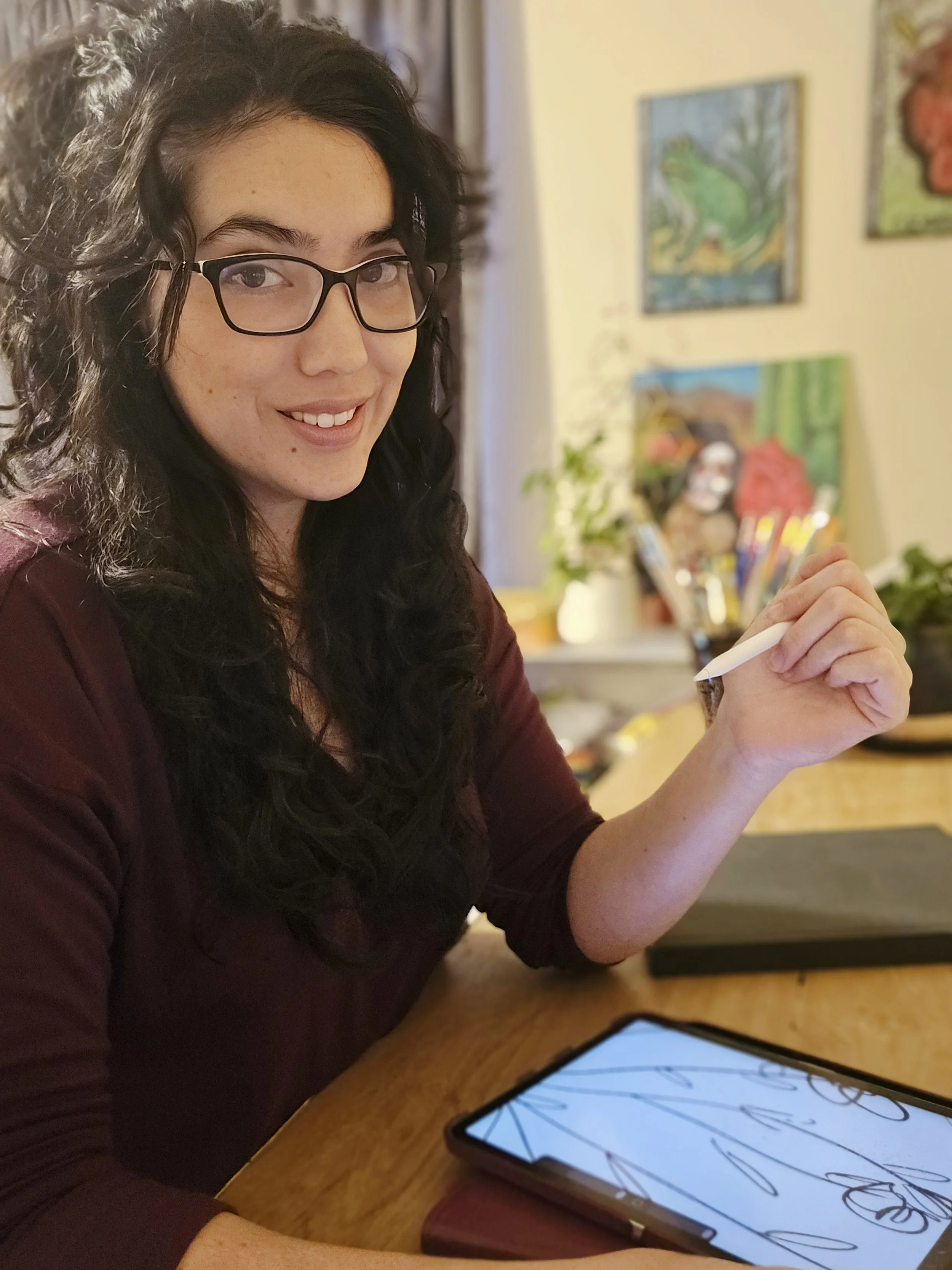 A woman with long curly dark hair, glasses, and light skin, sitting at a wooden table with a tablet displaying a drawing app, holding a stylus pen, in a room with colorful paintings and plants in the background.
