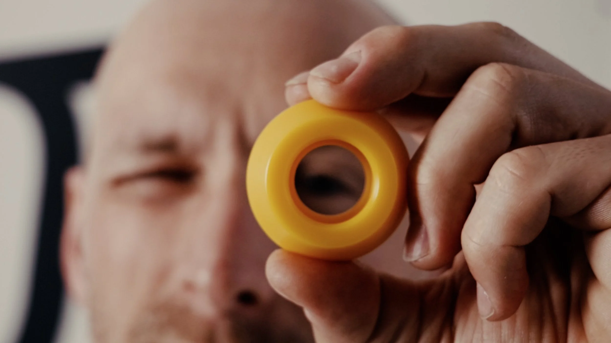 Mężczyzna trzyma w dłonie plastikowe kółko. Bardzo bliski kadr. Przez Środek kołka widać jego oko. Na pierwszym planie jest kółko które jest wyostrzone, mężczyzna na drugim planie jest rozmyty.