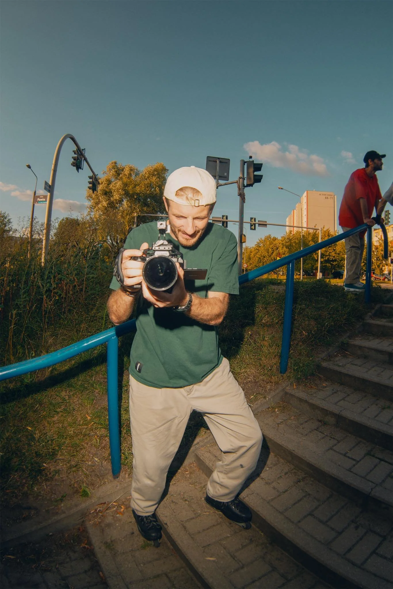 Mężczyzna ubrany w zieloną koszulę i beżowe spodnie, na rolkach, trzymający aparat, stojący na zewnątrz na schodach obok niebieskiej barierki, na tle drzew, sygnalizacji świetlnej i budynku w ciągu dnia.
