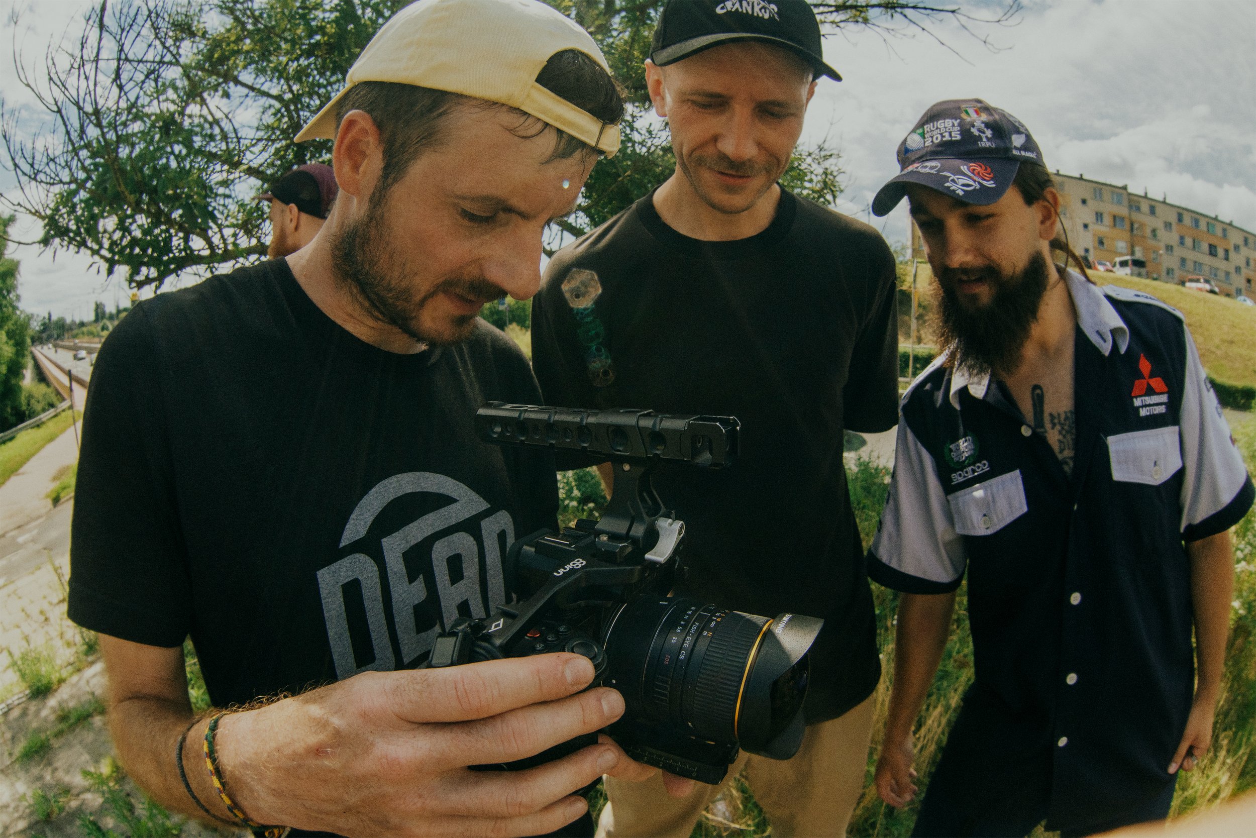 Trzech mężczyzn na zewnątrz patrzy na profesjonalną kamerę wideo, w tle widać porośnięte trawą wzgórze i budynki.