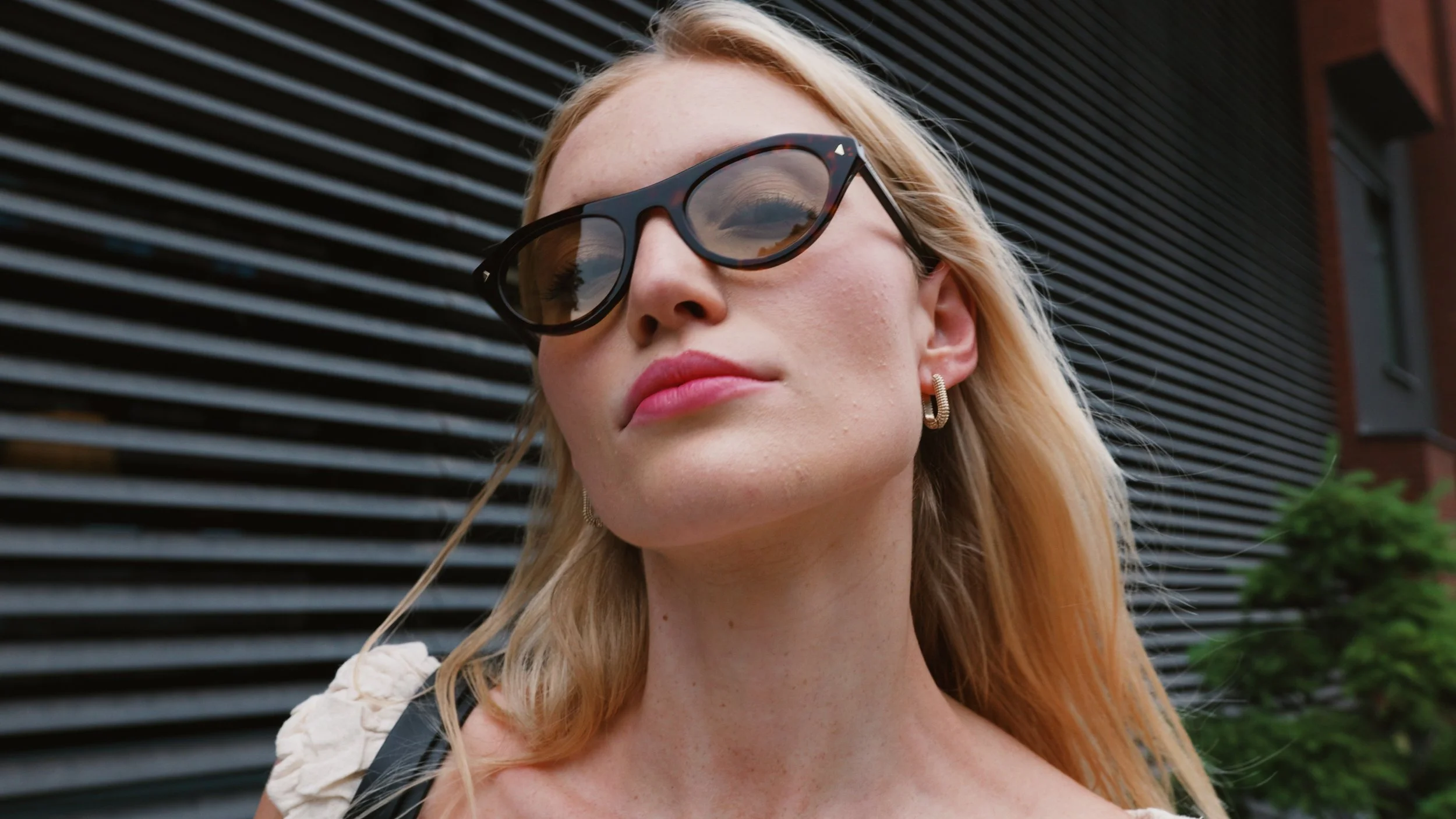 A woman with blonde hair wearing dark sunglasses, earrings, and pink lipstick, standing outside in front of a modern building with horizontal black panels and some greenery.