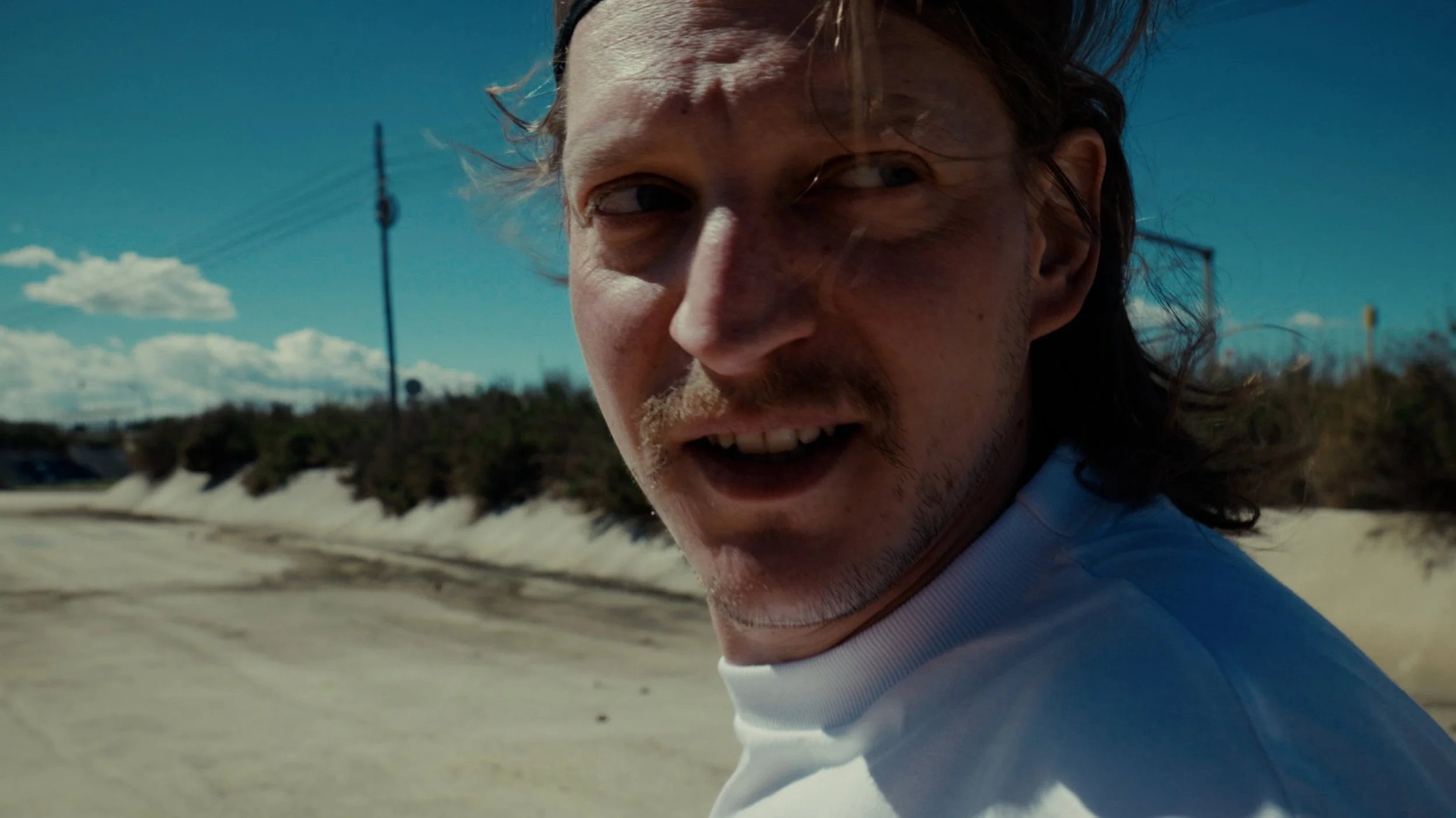 Close-up of a man with long hair, mustache, and beard, looking over his shoulder with a serious expression, outdoors on a sunny day with a blue sky and white clouds in the background.