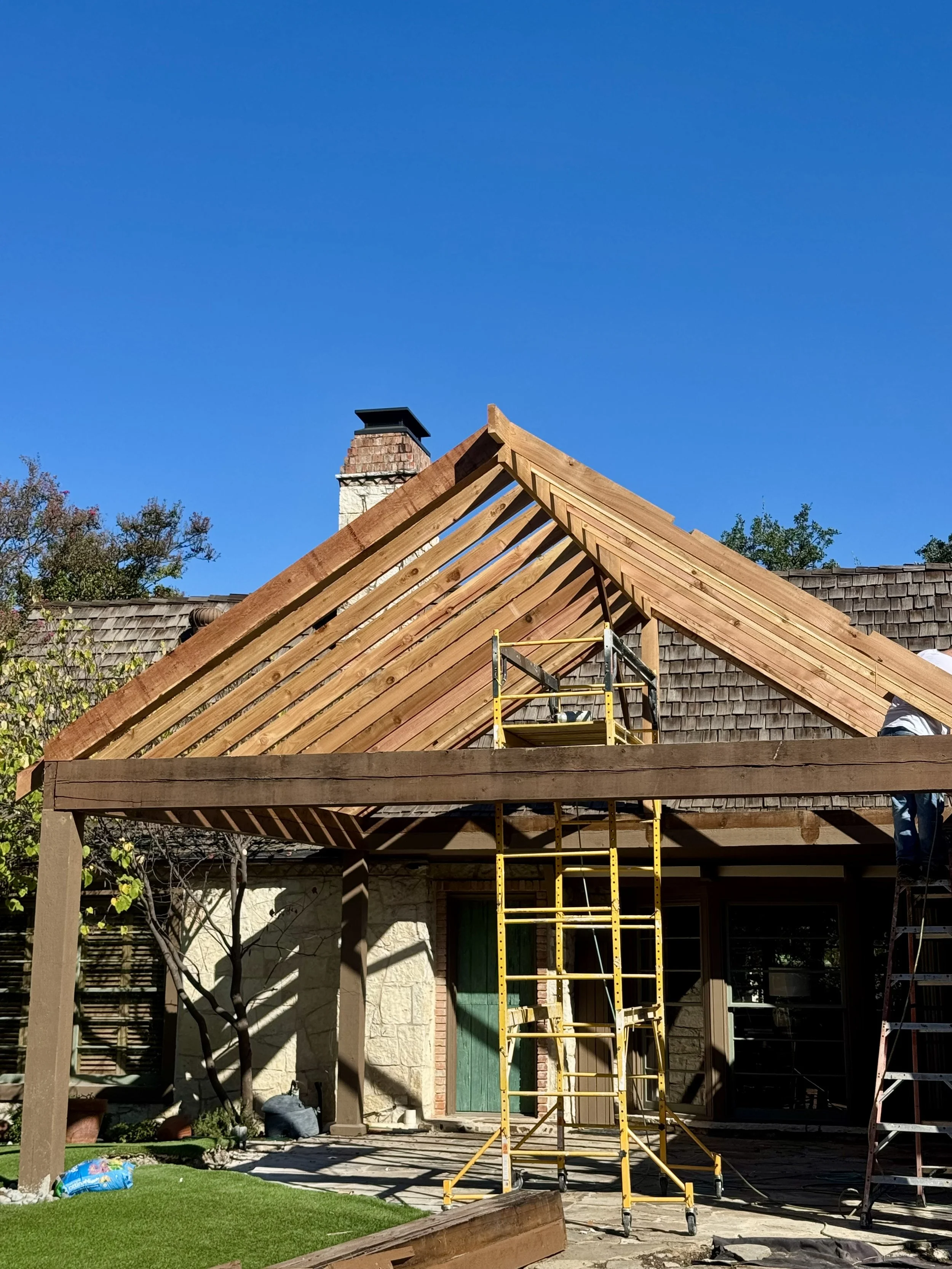 Patio structure under construction with new wooden roof framing, yellow scaffolding, and a ladder in front on a clear day.