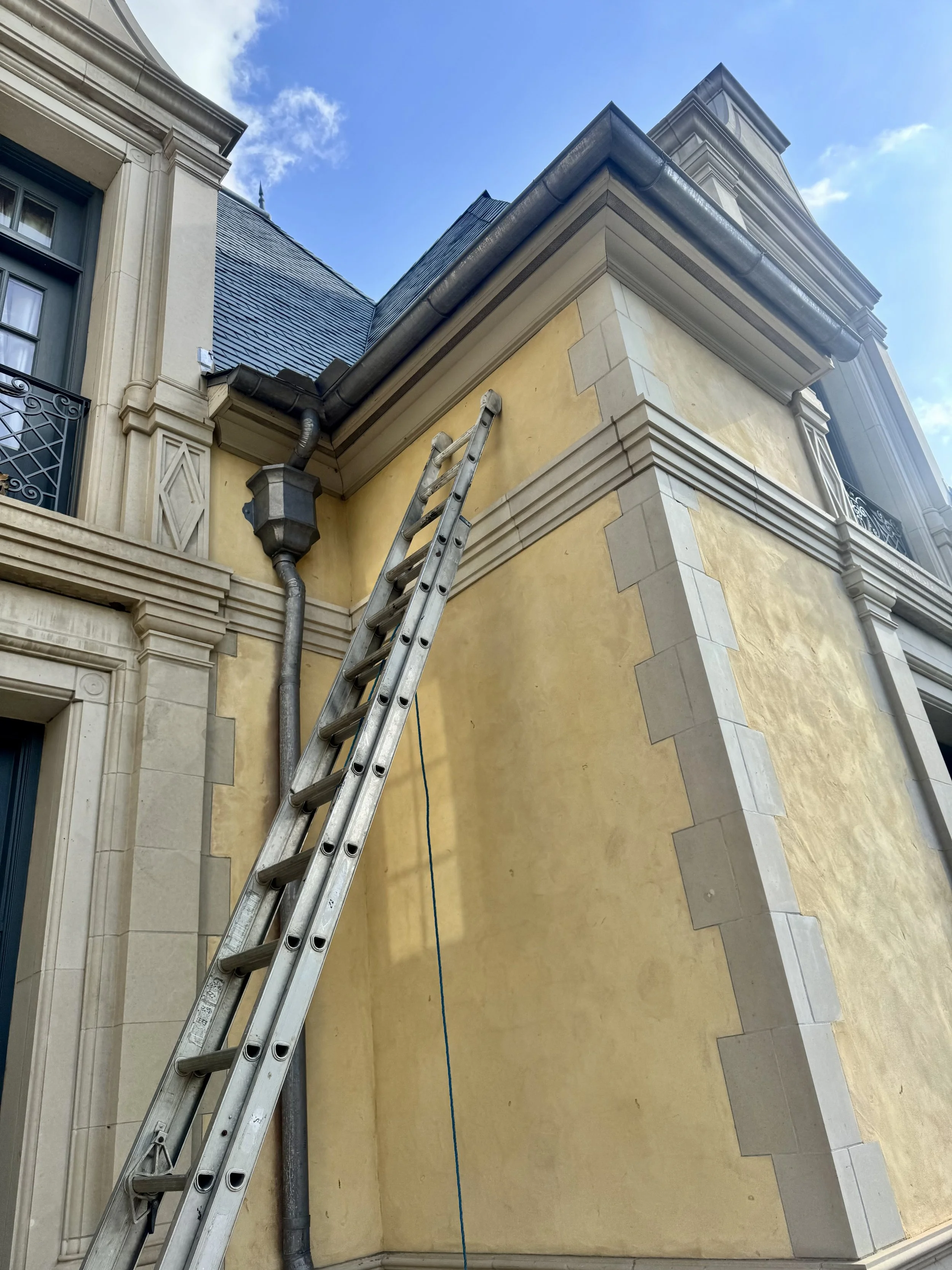 Ladder leaning against the exterior wall of a building under renovation or repair, with visible gutters, pipes, and architectural details.