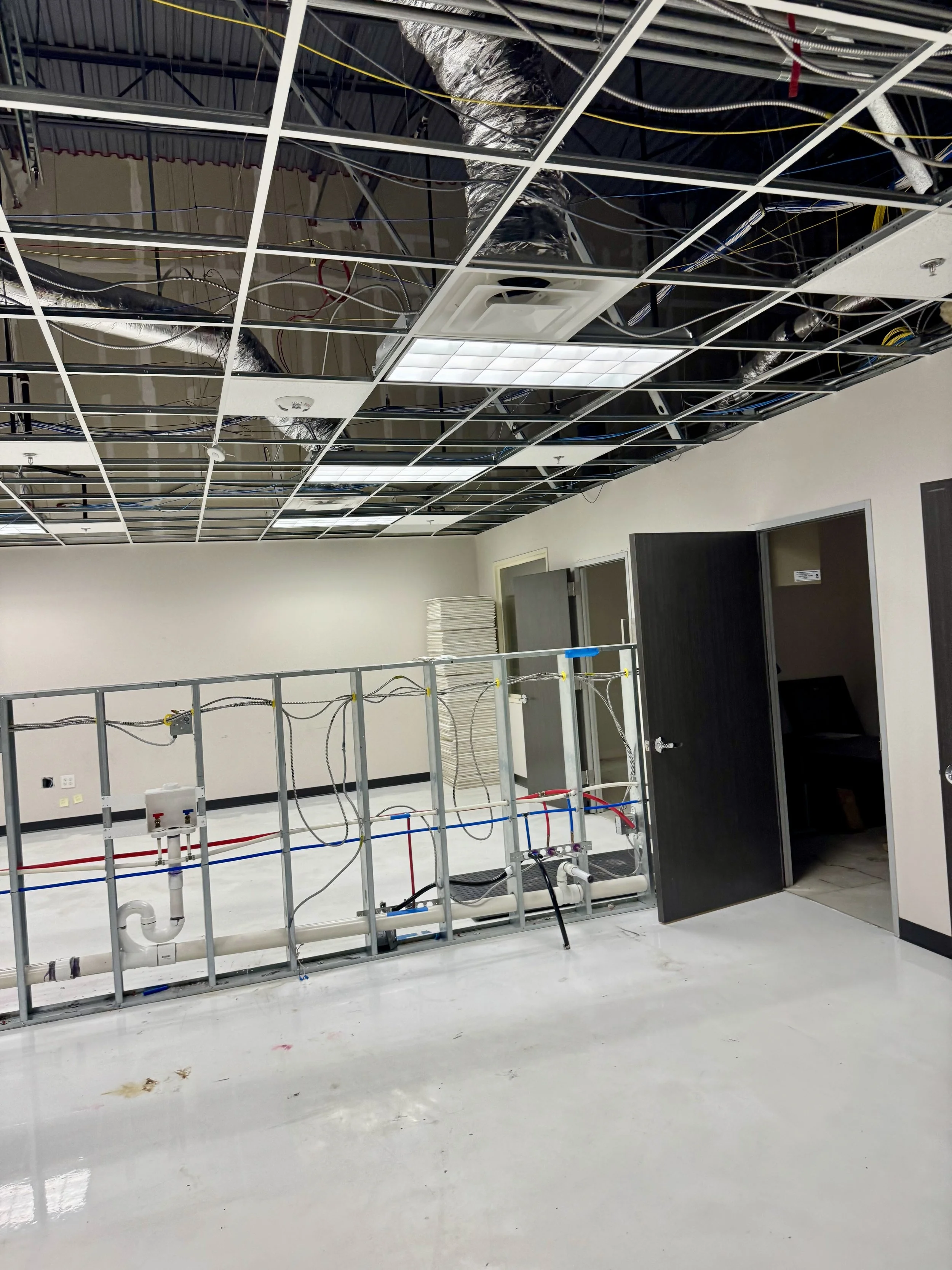 Office room under construction with exposed ceiling grid, electrical and plumbing components, and open doors.