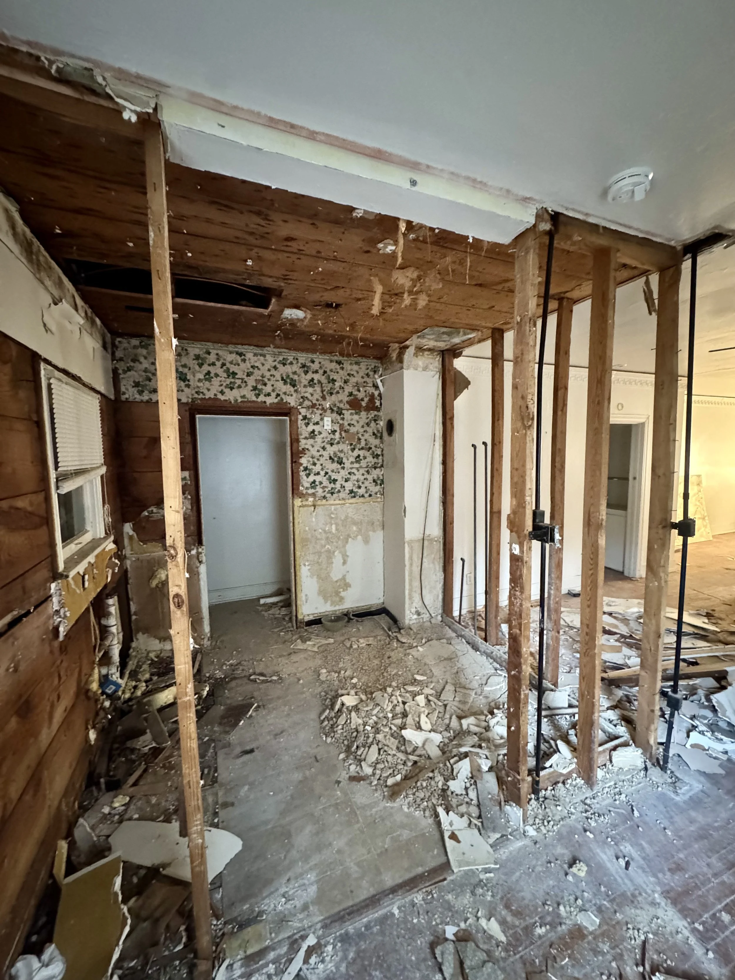 Room under renovation with exposed wooden studs, broken flooring, and partially removed walls, revealing insulation and plumbing.