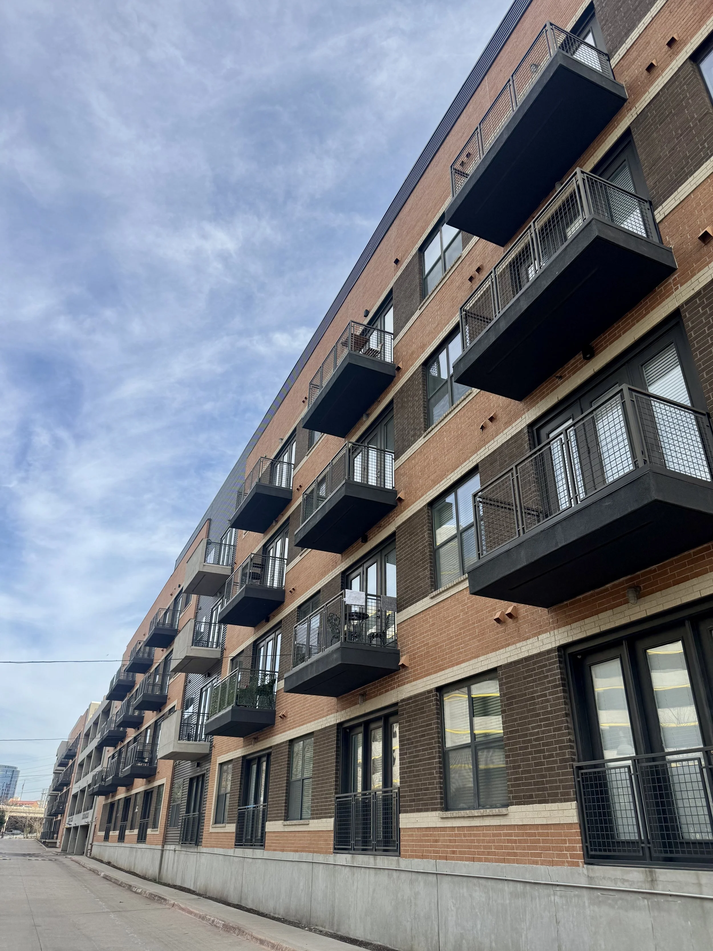 A multi-story urban apartment building with brick facade and black balconies under a partly cloudy sky.