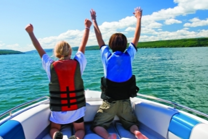 Two children on a boat with arms raised, enjoying a day on the water under a partly cloudy sky.