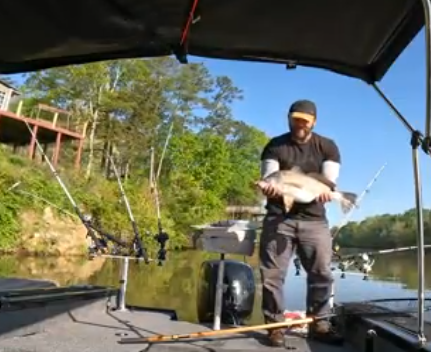 Fishing Guide Dustin Schmidt of Lake Weiss Destinations holding a fish on a boat during daytime with fishing rods and a scenic waterway background on Coosa River Alabama on Georgia border at Weiss Lake