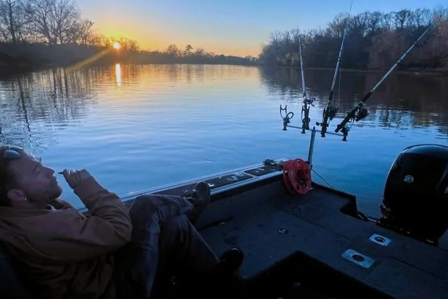 A person relaxing on a boat during sunset on a calm river with three fishing rods set up, and trees lining the riverbank in the background.