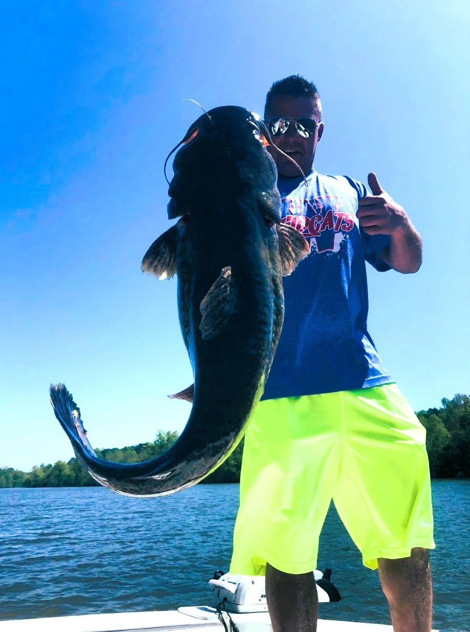Man on a boat holding a large fish he caught, smiling, wearing sunglasses, a gray T-shirt with red and white text, and bright yellow shorts, on a sunny day with blue sky and water in the background.