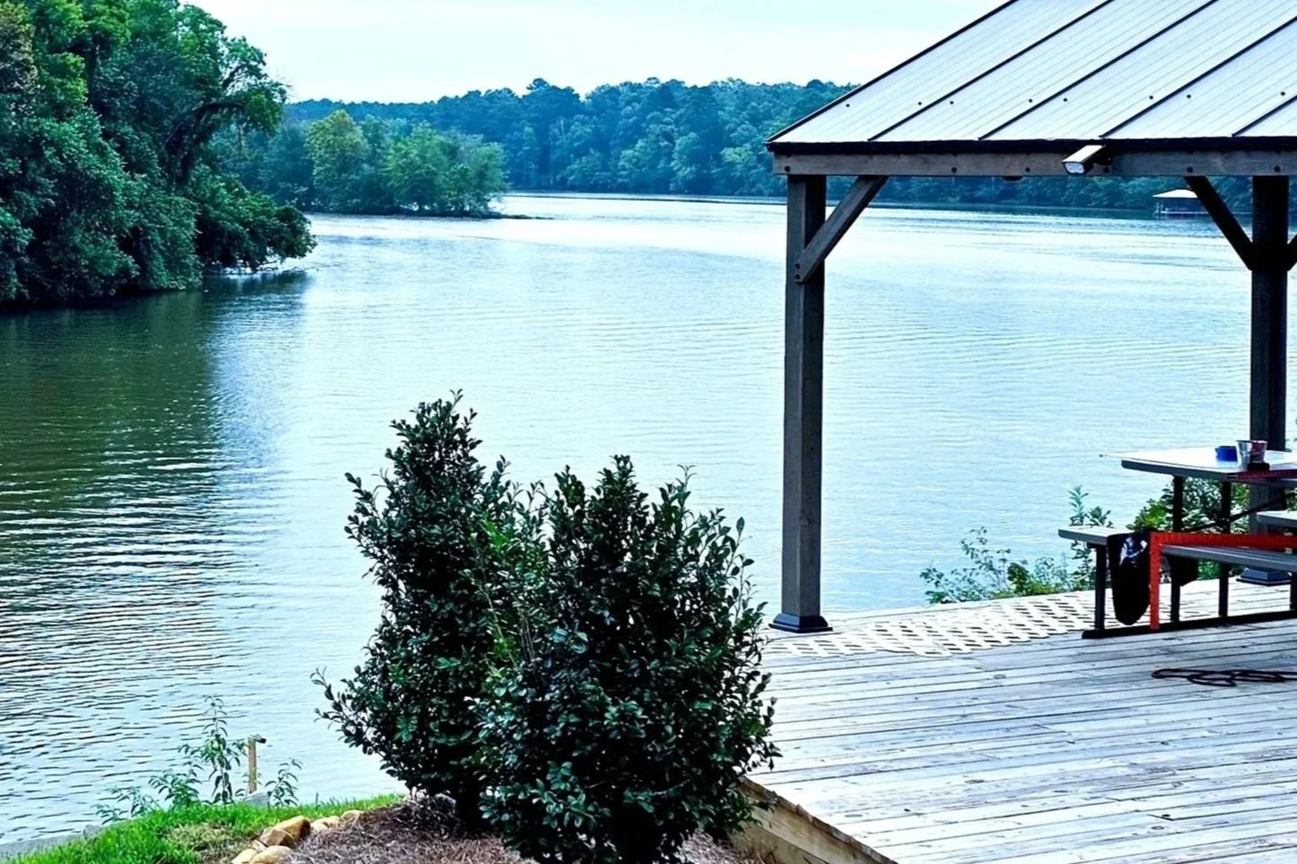 A lakeside view from a wooden deck with a small shrub in the foreground, a covered structure on the right, and a calm lake surrounded by lush green trees and distant hills.