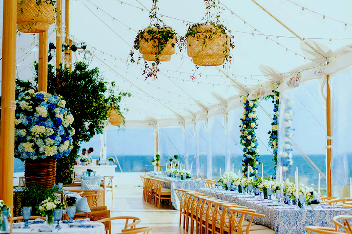 Decorated outdoor event space with floral arrangements, hanging potted plants, string lights, and ocean view.