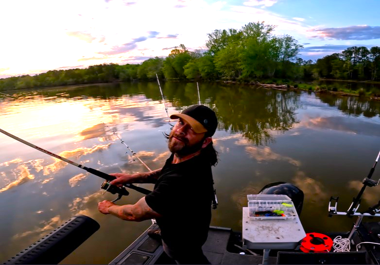 Fishing Guide Dustin Schmidt of Lake Weiss Destinations on a boat in a calm river during sunset, with a reflection of clouds and trees on the water, and fishing gear and tackle box nearby  on Coosa River Alabama on Georgia border at Weiss Lake