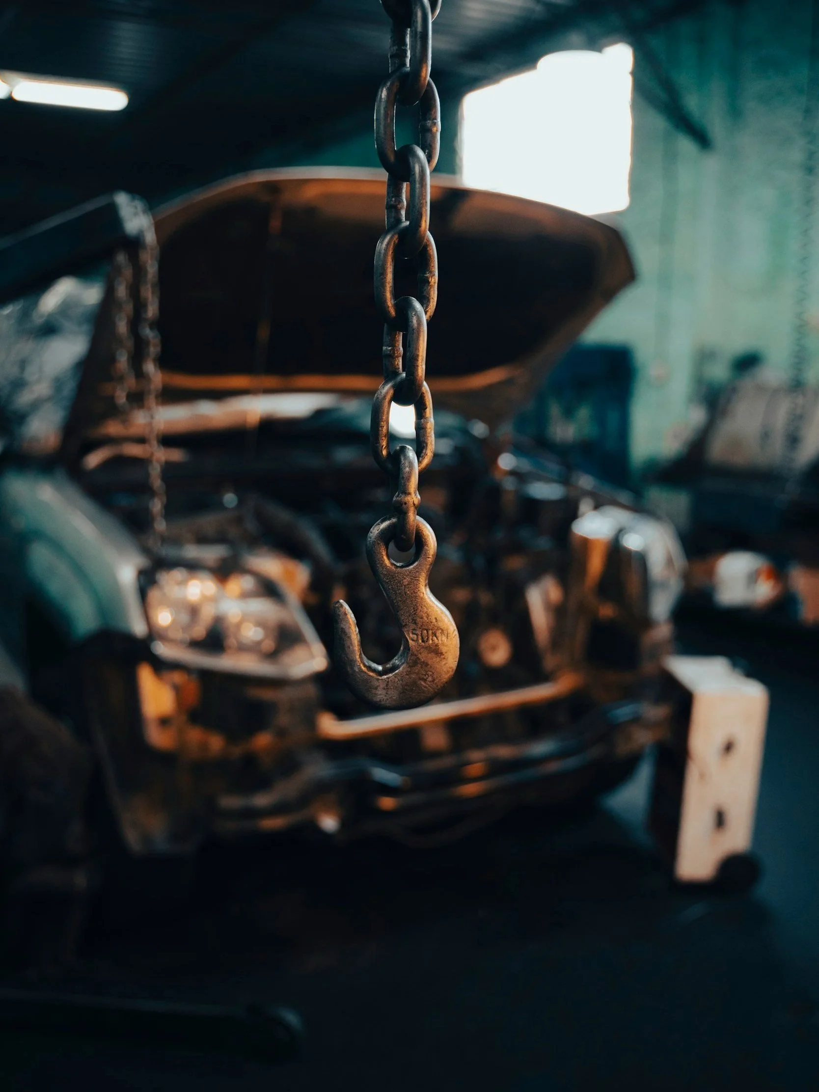 Close-up of a metallic hook hanging from a chain in an automotive repair garage, with a car undergoing repair in the background.