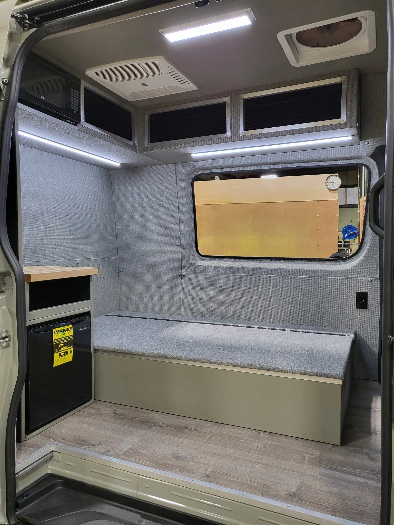 Interior of a converted camper van with a small fridge, a countertop, a cushioned seating area, a large window, and overhead storage compartments.