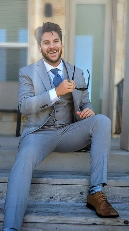 A man in a light gray suit sitting on wooden steps outside, smiling while holding sunglasses.