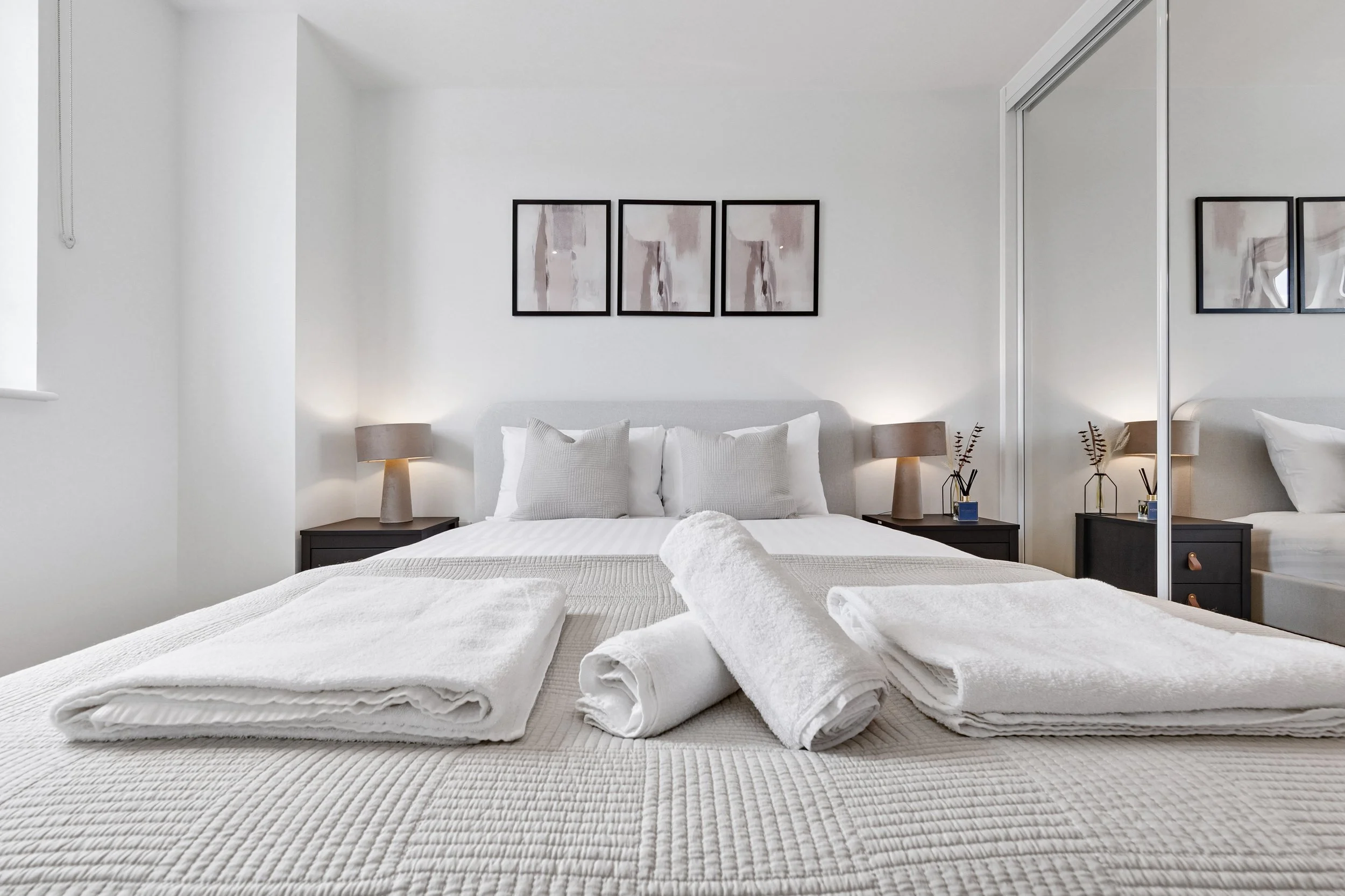 Neat bedroom with white bedding, three pillows, framed artwork above bed, two bedside lamps, mirrored closet door, and folded towels on bed.
