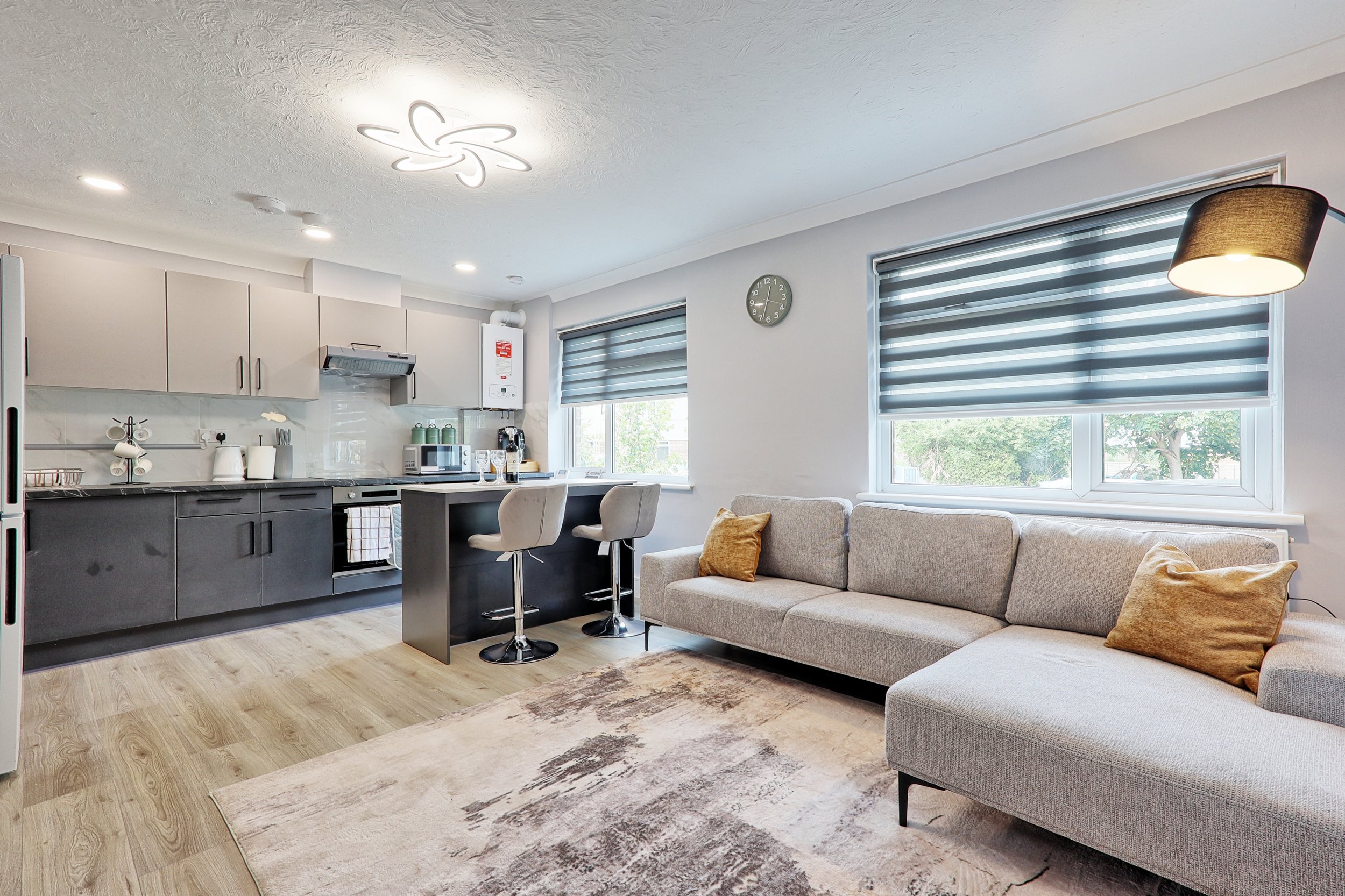 Open-plan living room and kitchen with gray and beige furniture, wooden flooring, and modern decor including a gray sofa with brown pillows, kitchen cabinets, a breakfast bar with two beige chairs, and large windows with back-and-white striped blinds.