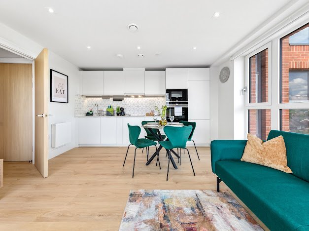 Open-plan kitchen and living room with white cabinets, wooden flooring, a dining table with green chairs, a teal sofa with pillows, a colorful rug, and large windows with a view of neighboring buildings.