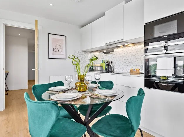 Modern kitchen with white cabinets, a round dining table with teal chairs, and decorative green plants in a gold vase.