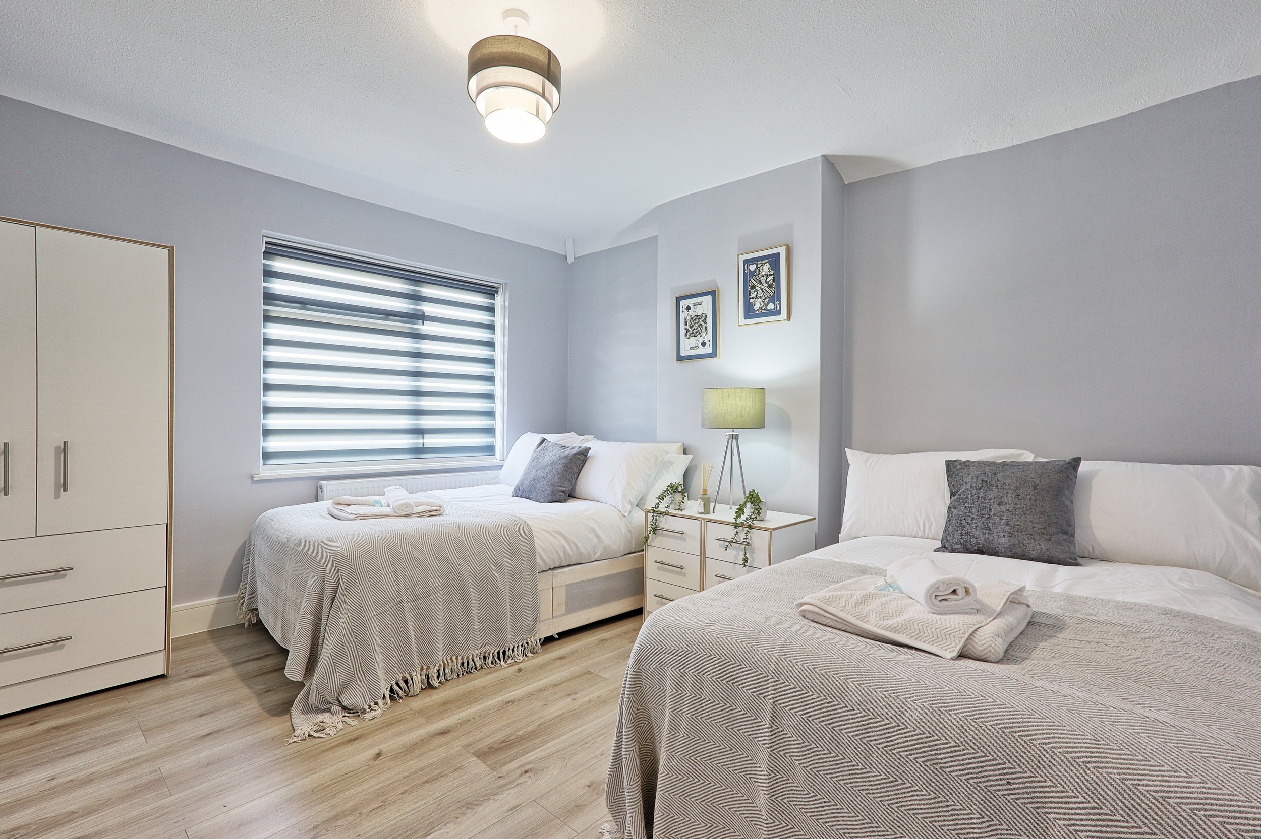 A bedroom with two neatly made beds with light grey and white bedding, a nightstand with a lamp and decor, a window with horizontal blinds, a wardrobe, and wall art, all in a modern, minimalist style.
