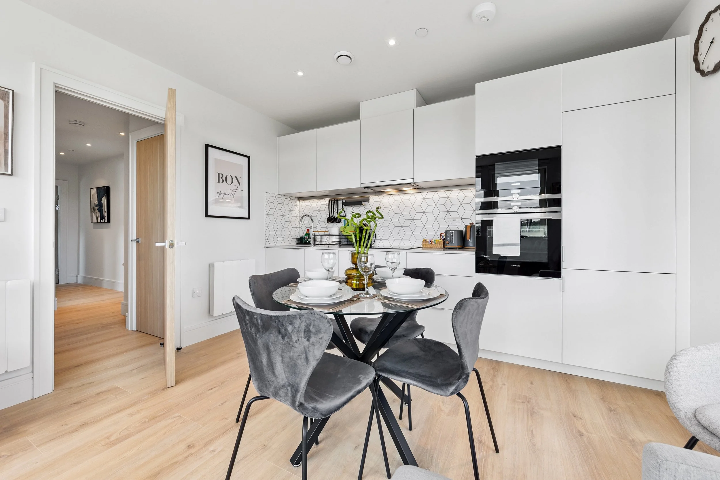 Modern kitchen and dining area with white cabinets, black oven, gray chairs, round glass table, wooden flooring, decorative items, and artwork on the wall.