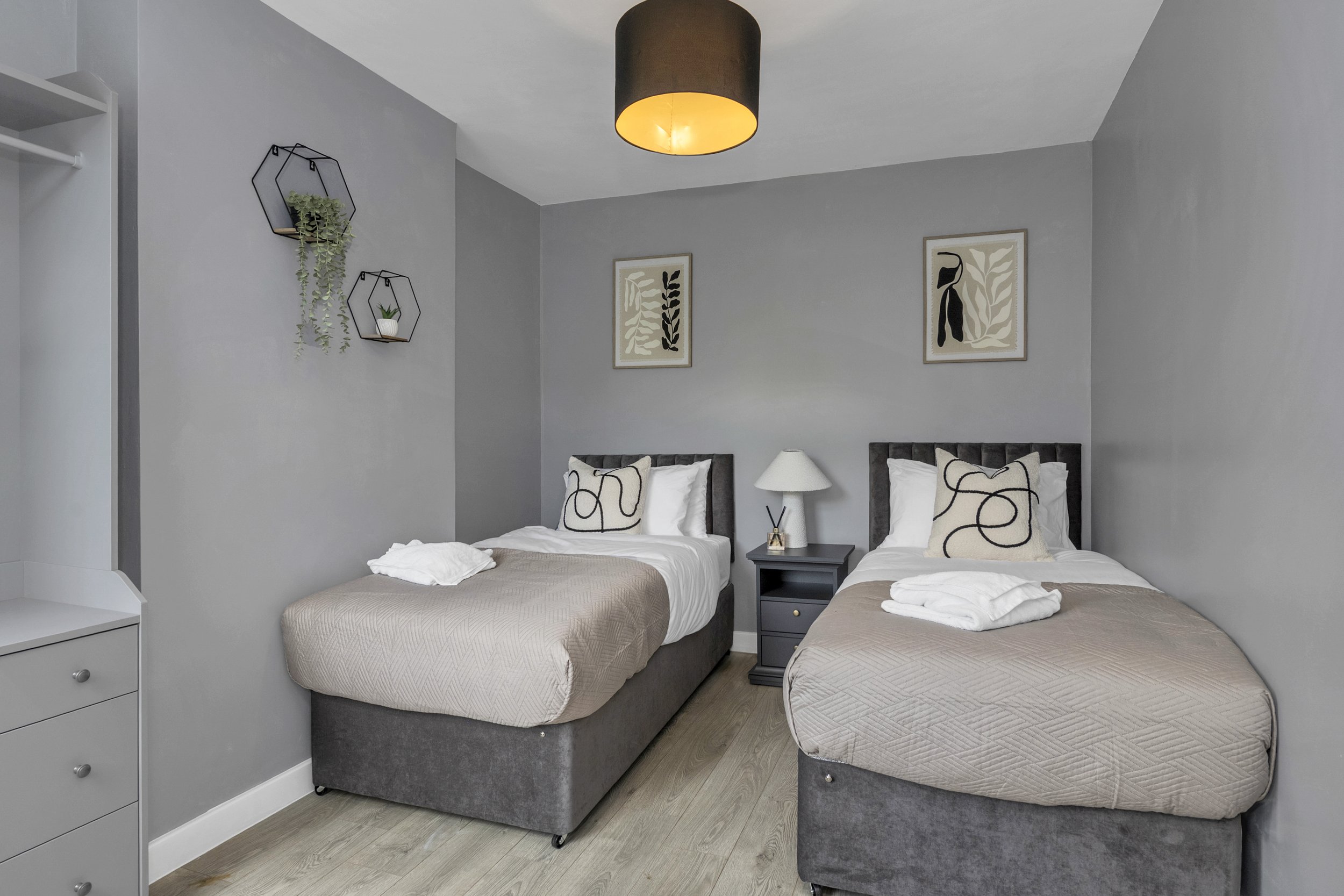 Bedroom with two single beds with beige quilts, white sheets, decorative pillows, and dark gray headboards, a small black nightstand with a white lamp in the middle, gray walls, framed botanical art, and wall-mounted planters.