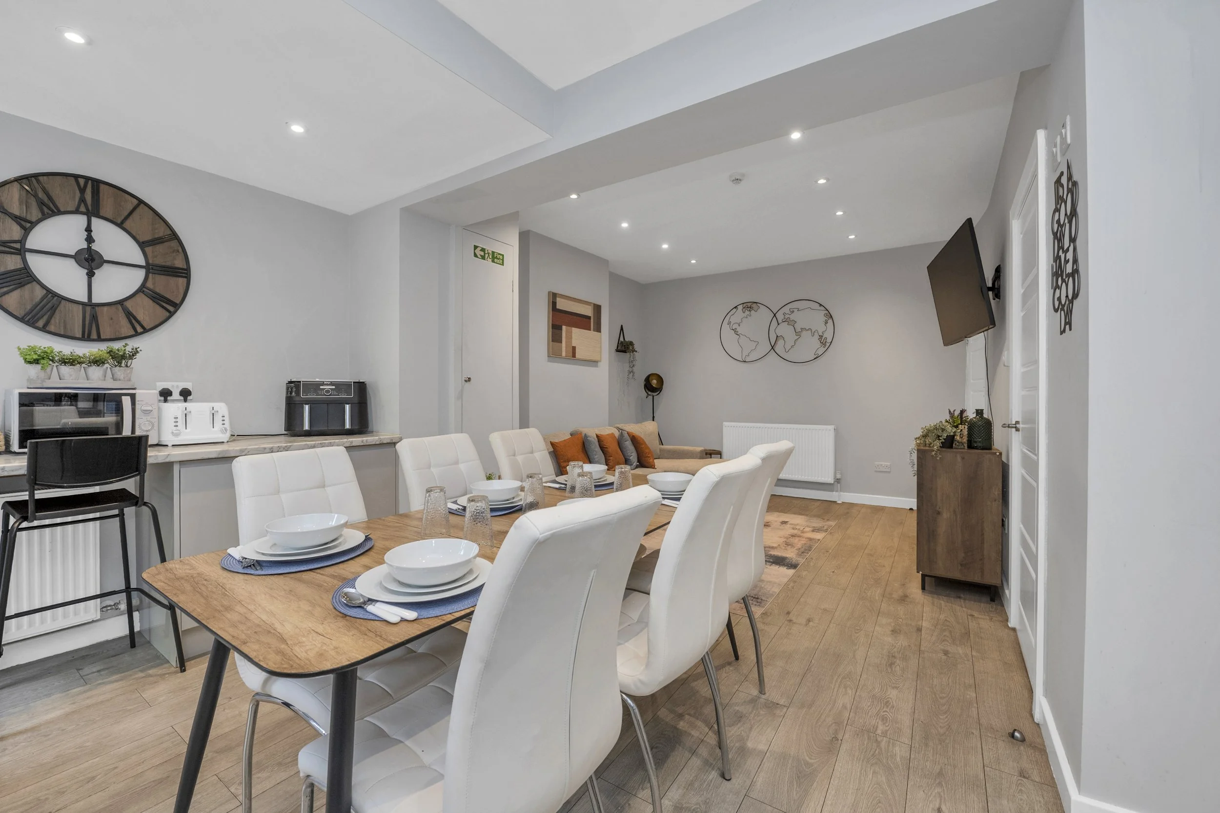 Modern dining and living room with white chairs around a wooden table set with plates and glasses, a wall clock, TV, world map art, and wooden flooring.
