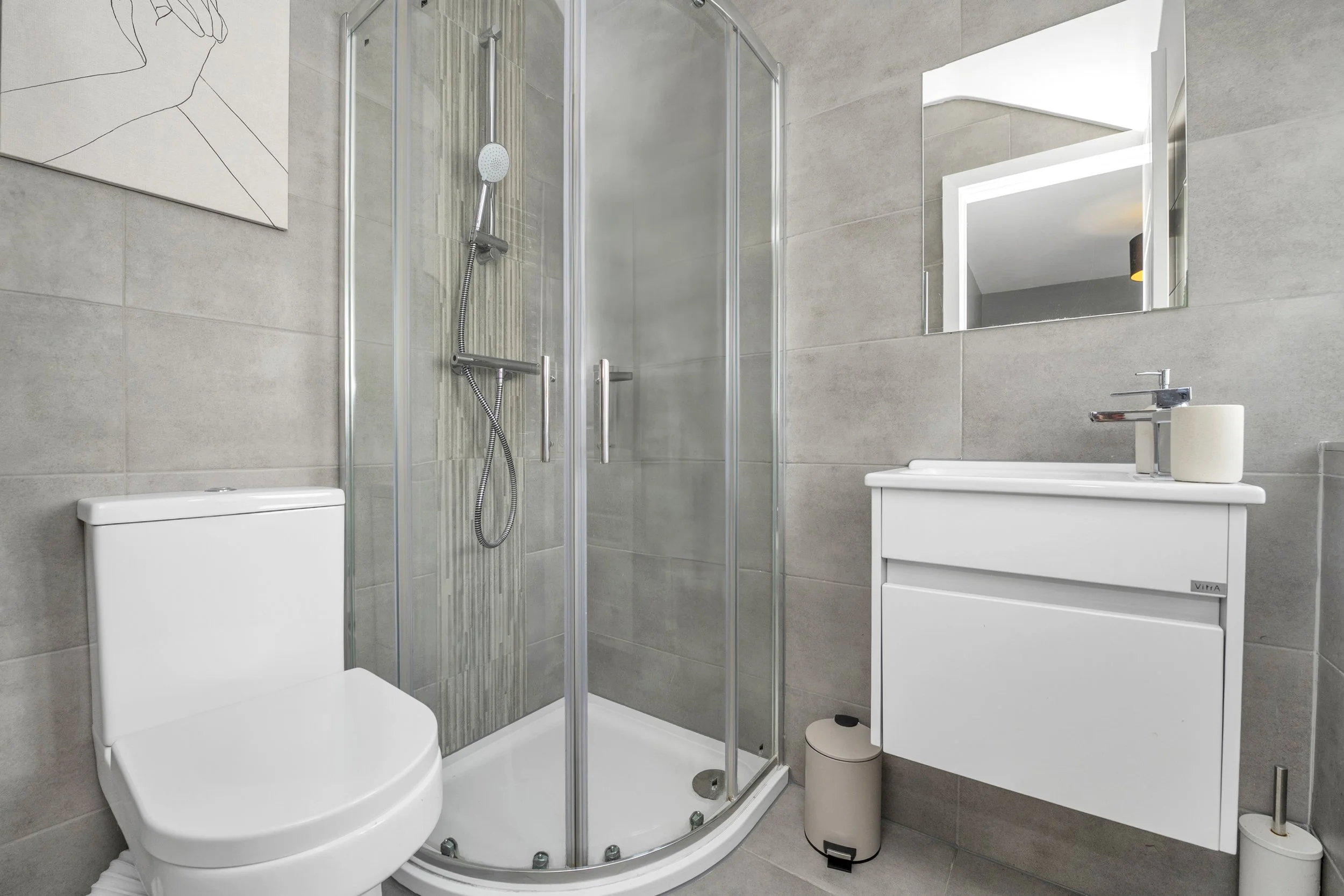 Modern bathroom with a toilet, a shower with glass doors, a white vanity with a sink, and a mirror on the wall. The walls and floor are covered with beige tiles.