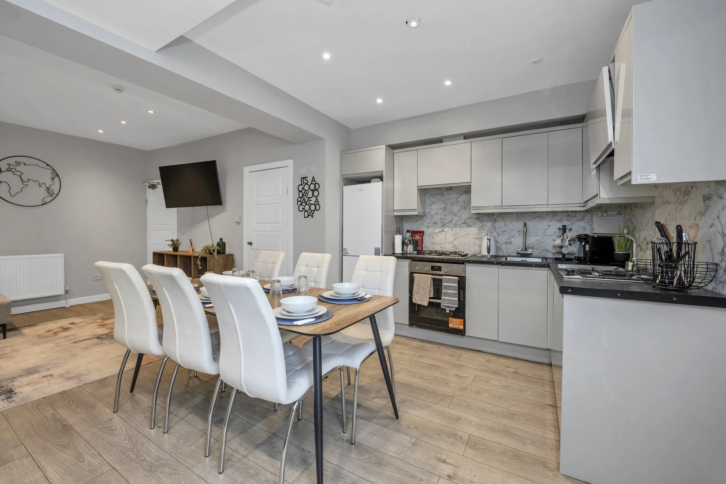 Modern kitchen and dining area with a wooden table set for six, white upholstered chairs, gray cabinetry, marble backsplash, and various appliances.