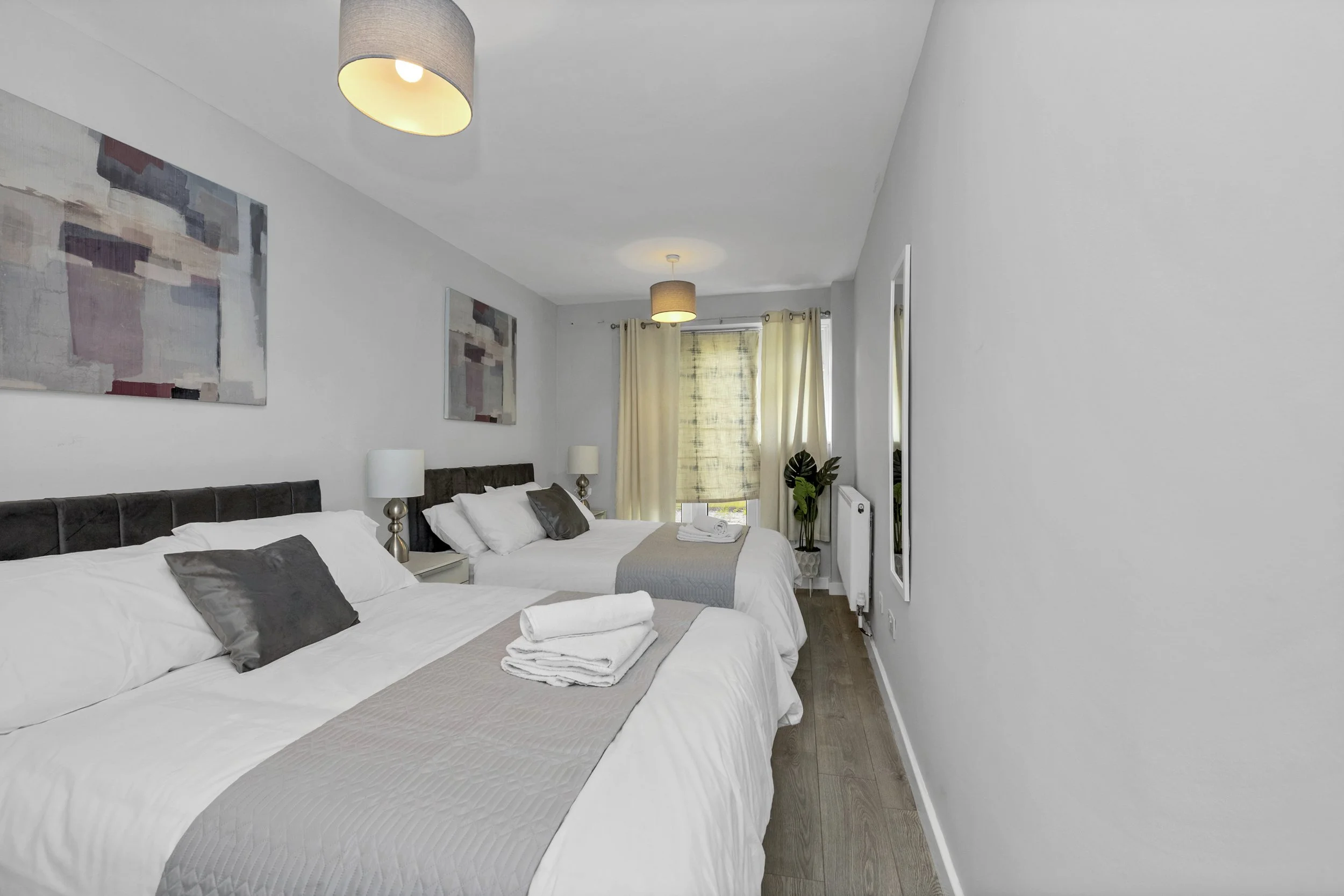 A modern bedroom with two beds, white bedding, dark pillows, beige runners, and small bedside tables with lamps. Artwork hangs on the white walls, and a window with yellow curtains and a roller blind is at the end of the room. Two towels are folded on each bed, and a potted plant is near the window.
